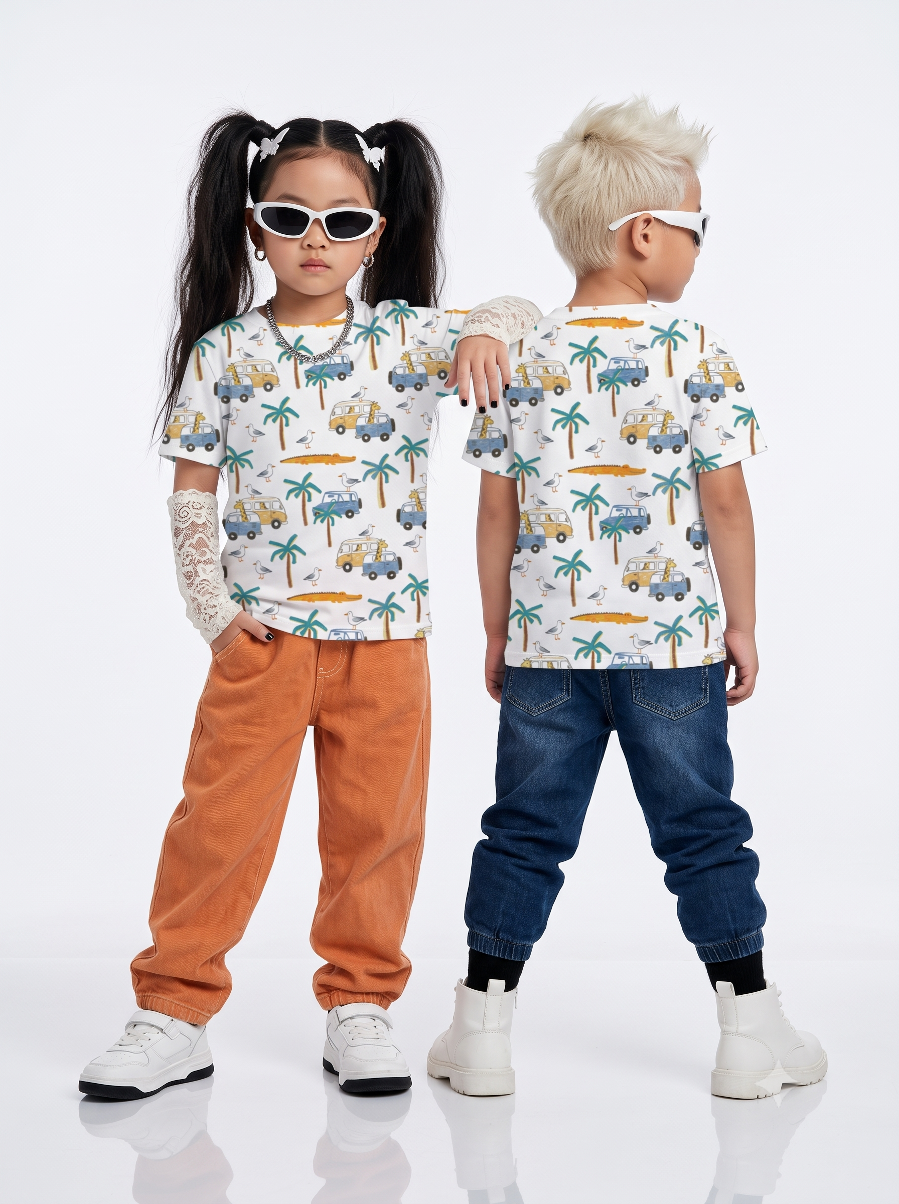 Two children modeling matching short-sleeve t-shirts with a tropical truck and palm tree print, styled with sunglasses, orange pants, blue jeans, and white sneakers against a white studio background.